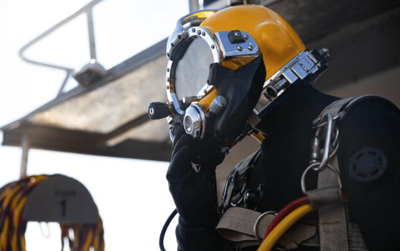 Screenshot 2026-04-07 172403 - N9BO℠ | Global Underwater Services Ltd A person wearing a yellow diving helmet and black wetsuit prepares for a dive, holding their helmet, with safety harnesses and cables attached. A boat structure and coiled cords are visible in the background.