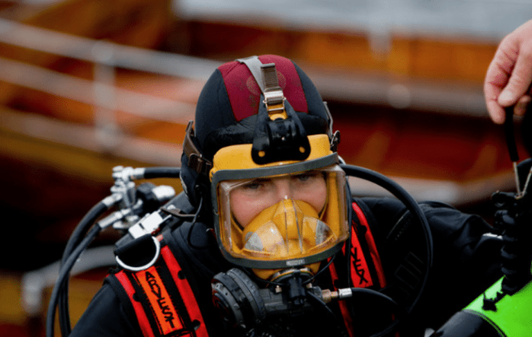 A person wearing a black and red wetsuit and a full-face scuba diving mask with attached breathing apparatus, preparing for a dive. The blurred background appears to show wooden structures or boats.