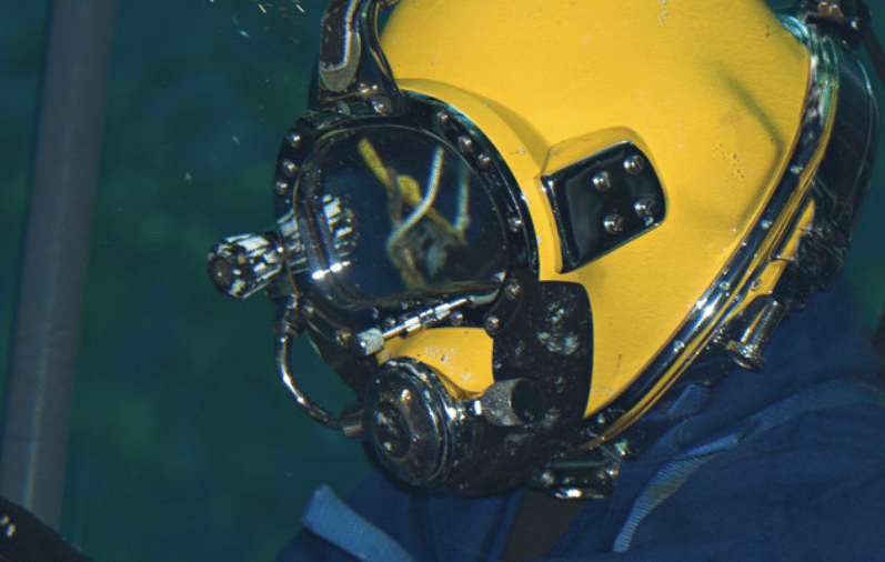 Screenshot 2026-04-07 172501 - N9BO℠ | Global Underwater Services Ltd A close-up of a person underwater wearing a yellow commercial diving helmet with a clear visor and metal fittings, dressed in a blue suit.