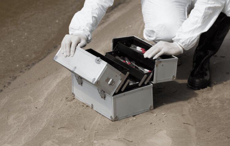 A person wearing white protective gloves and a suit kneels on sandy ground, opening a metal case filled with vials and scientific equipment near the edge of water.