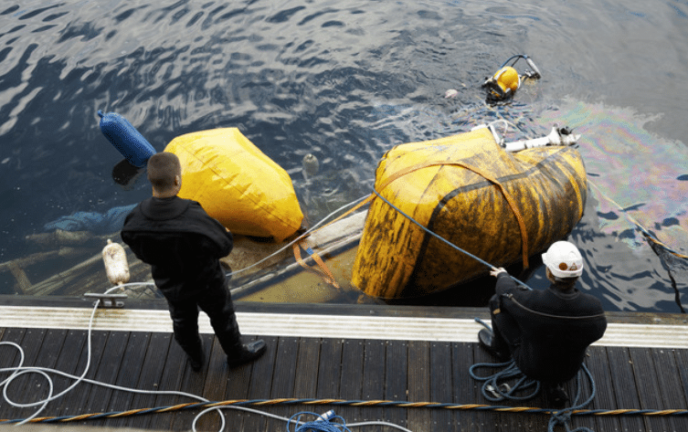 Screenshot 2026-04-07 173646 - N9BO℠ | Global Underwater Services Ltd Two people stand by the water’s edge, overseeing a diver and large yellow flotation bags partially submerged, possibly involved in a salvage or recovery operation. One person holds ropes attached to the bags.