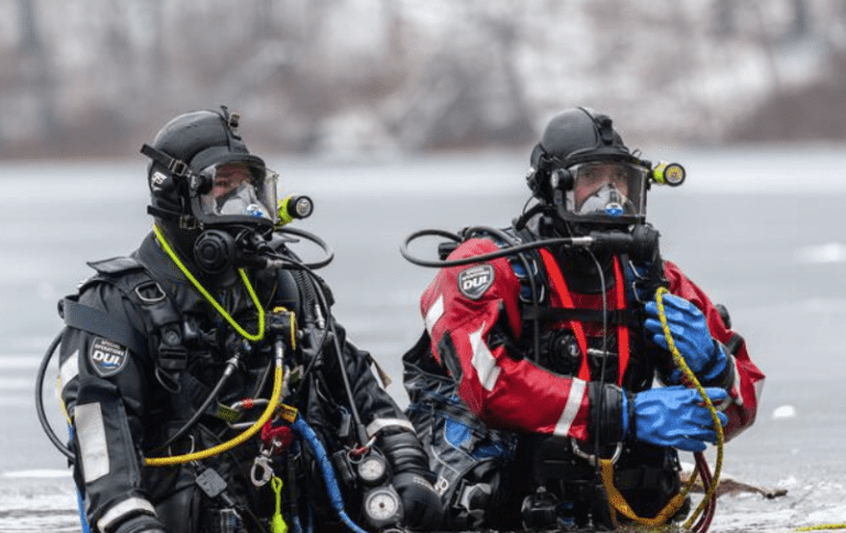 Two scuba divers in full kit, one in black and one in red, kneel at the edge of an icy body of water. Both wear masks and gloves, and are connected by yellow cords, with a snowy background visible.