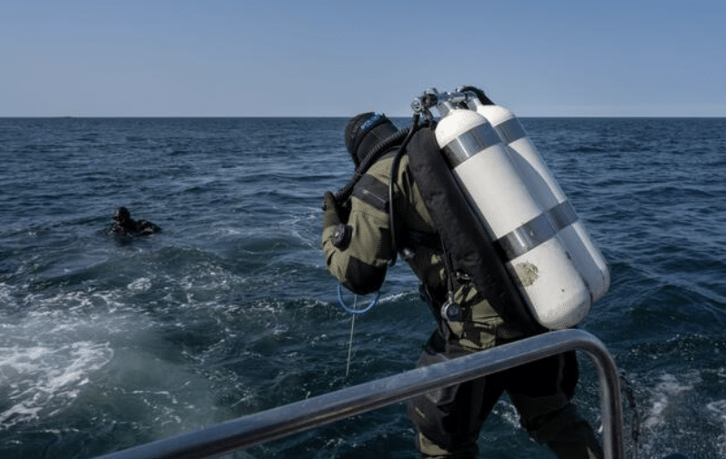 A diver wearing scuba gear prepares to enter the sea from a boat, while another diver is already in the water nearby under a clear blue sky.
