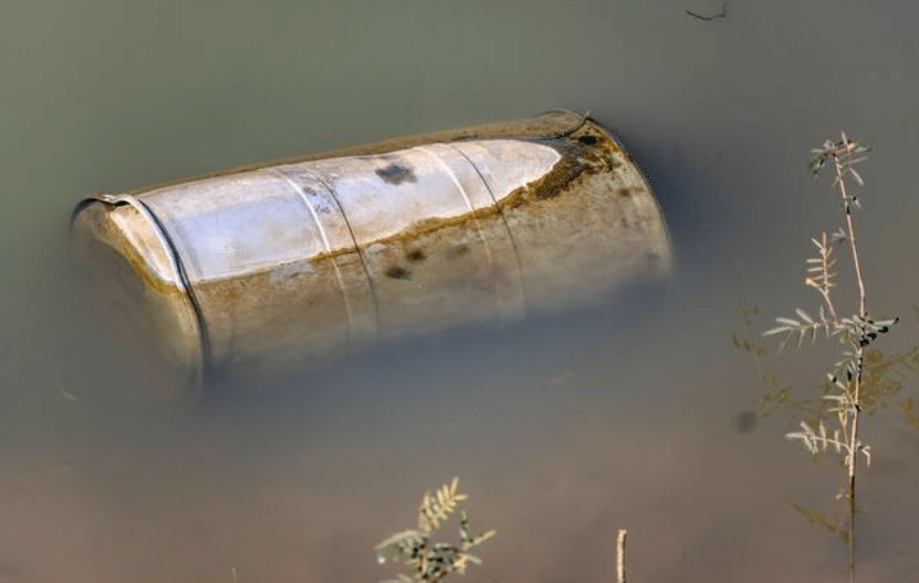 Screenshot 2026-04-07 175602 - N9BO℠ | Global Underwater Services Ltd A rusty metal barrel partially submerged in murky water, with some algae and plant growth visible around its edges. A few small plants are growing in the foreground near the water’s surface.