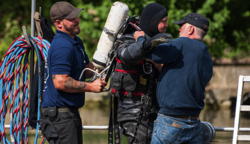 Screenshot 2026-04-07 175623 - N9BO℠ | Global Underwater Services Ltd Three men assist a diver in a wetsuit and scuba gear near water; one man holds cables, another helps the diver with equipment. Green foliage is visible in the background.