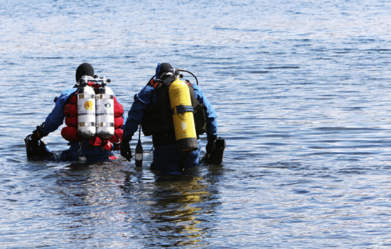 Two scuba divers in full kit wade into the water, each carrying oxygen cylinders on their backs, preparing to dive in a calm body of water.