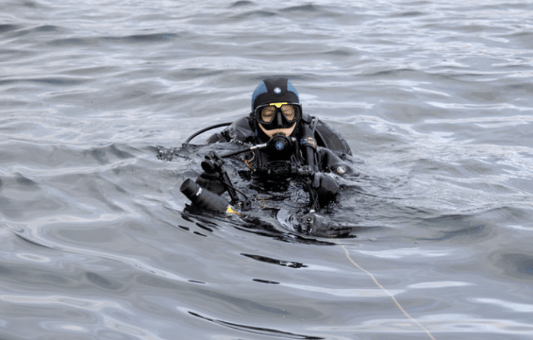 A scuba diver in full kit, including a mask and wetsuit, floats on the surface of calm, rippling water, holding an object and connected to a safety line.