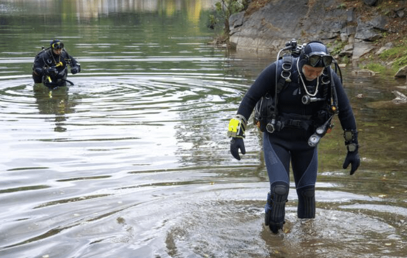 Two scuba divers in full kit walk out of a calm body of water towards a rocky shore, with one diver closer to the camera and the other further behind.