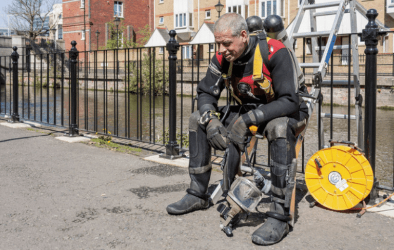 A man in scuba diving gear sits on a bench near a river, looking tired. He is holding his gloves and equipment, with a coiled yellow rope and ladder nearby. Buildings and a metal fence are in the background.