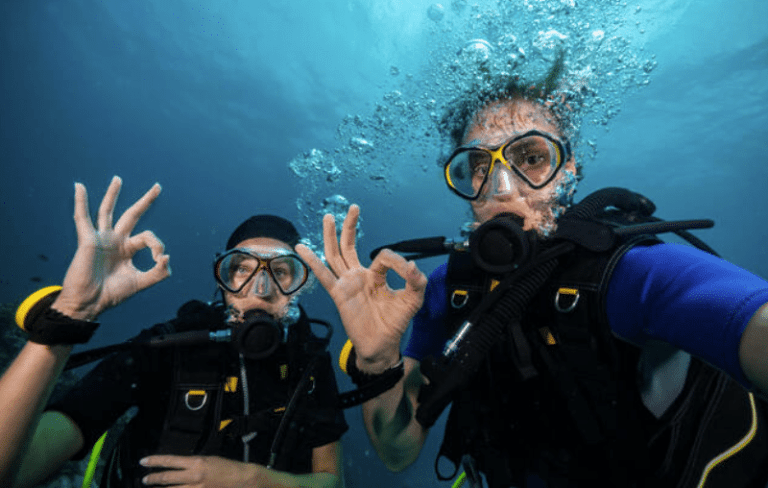 Two scuba divers underwater wearing masks and wetsuits give the “OK” hand signal to the camera, with bubbles rising above their heads and clear blue water in the background.