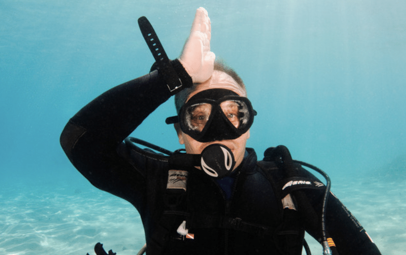 A scuba diver underwater makes a hand gesture by placing the side of his hand on his forehead, signalling shark while wearing diving kit and a mask. Sunlight filters through the clear blue water.