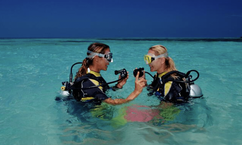 Two women wearing scuba gear stand in clear, shallow turquoise water under a blue sky. They face each other, smiling and talking, possibly preparing for a dive.