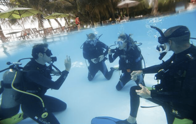 Four people wearing scuba gear kneel underwater in a swimming pool, appearing to take part in a scuba diving lesson. Palm trees, sun umbrellas, and loungers can be seen around the pool above the water.