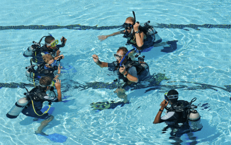 A group of people in scuba gear practise diving skills with an instructor in a clear swimming pool, communicating using hand signals underwater.