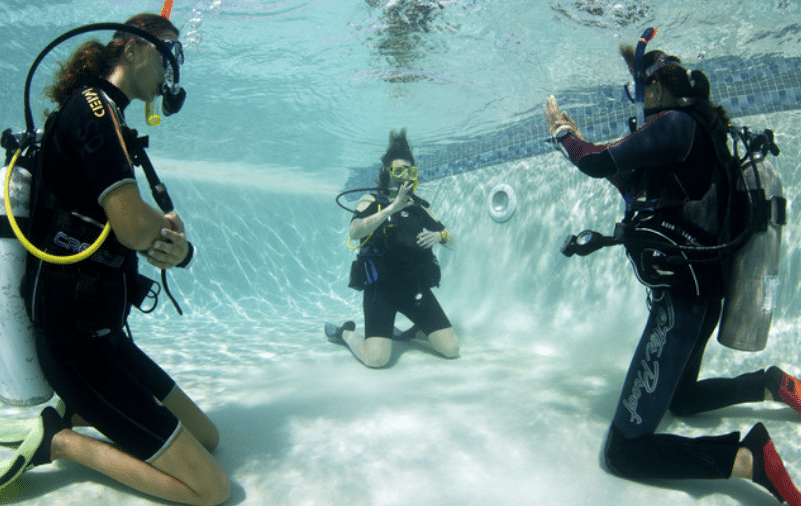 Three scuba divers in wetsuits and kit kneel on the bottom of a swimming pool. One appears to be instructing the others, using hand signals, while another kneels with hands together, and the third observes closely.