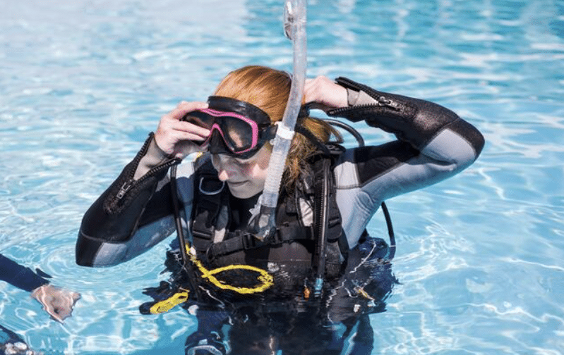 A person wearing scuba diving gear and a black wetsuit adjusts their diving mask whilst standing in clear blue water, preparing for a dive.
