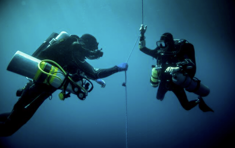 Screenshot 2026-04-07 201200 - N9BO℠ | Global Underwater Services Ltd Two scuba divers in full kit hold onto a guide rope underwater, surrounded by deep blue water, as they communicate and maintain their position during a dive.