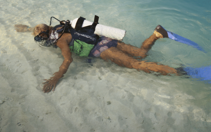 A scuba diver wearing fins and a cylinder swims face down in clear, shallow water over a sandy seabed.