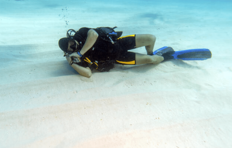 A scuba diver in black kit and blue fins lies on the sandy seabed, checking a device on their wrist underwater.