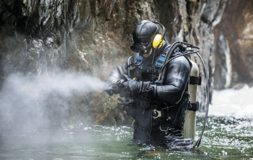 A diver wearing a wetsuit, helmet, and yellow headphones uses a high-pressure water hose whilst standing in a rocky, shallow water area. Mist and water spray surround the diver.
