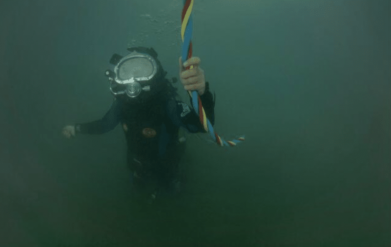 Screenshot 2026-04-07 204802 - N9BO℠ | Global Underwater Services Ltd A diver in a mask and wetsuit underwater holds onto a colourful, twisted rope. The visibility is low, and the water appears murky.