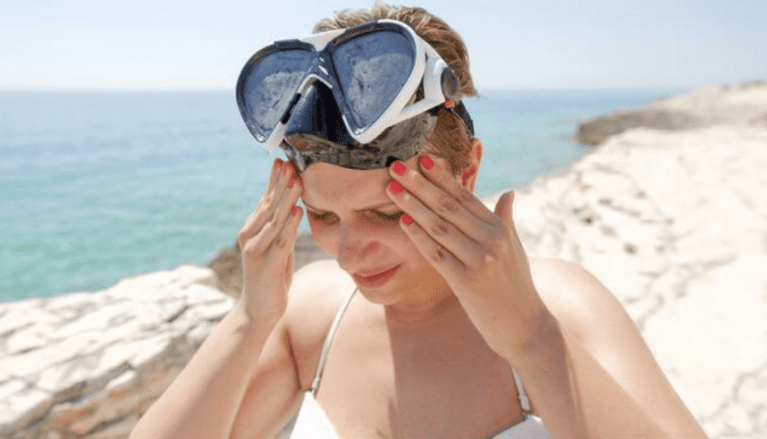 A woman with red nail varnish adjusts her large snorkelling mask on her forehead whilst standing near rocky white cliffs by a calm blue sea under a clear sky.