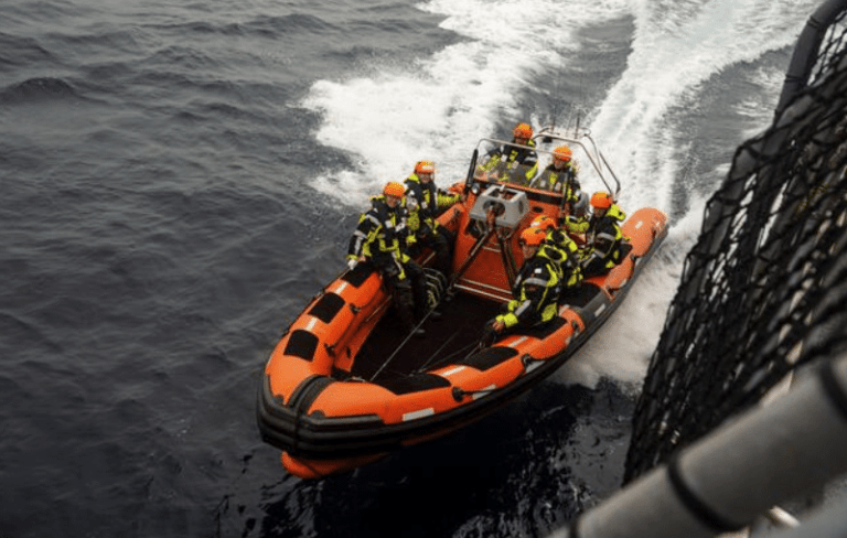A group of people wearing helmets and life jackets ride an orange inflatable rescue boat on the sea, creating a white trail in the water next to a larger vessel.