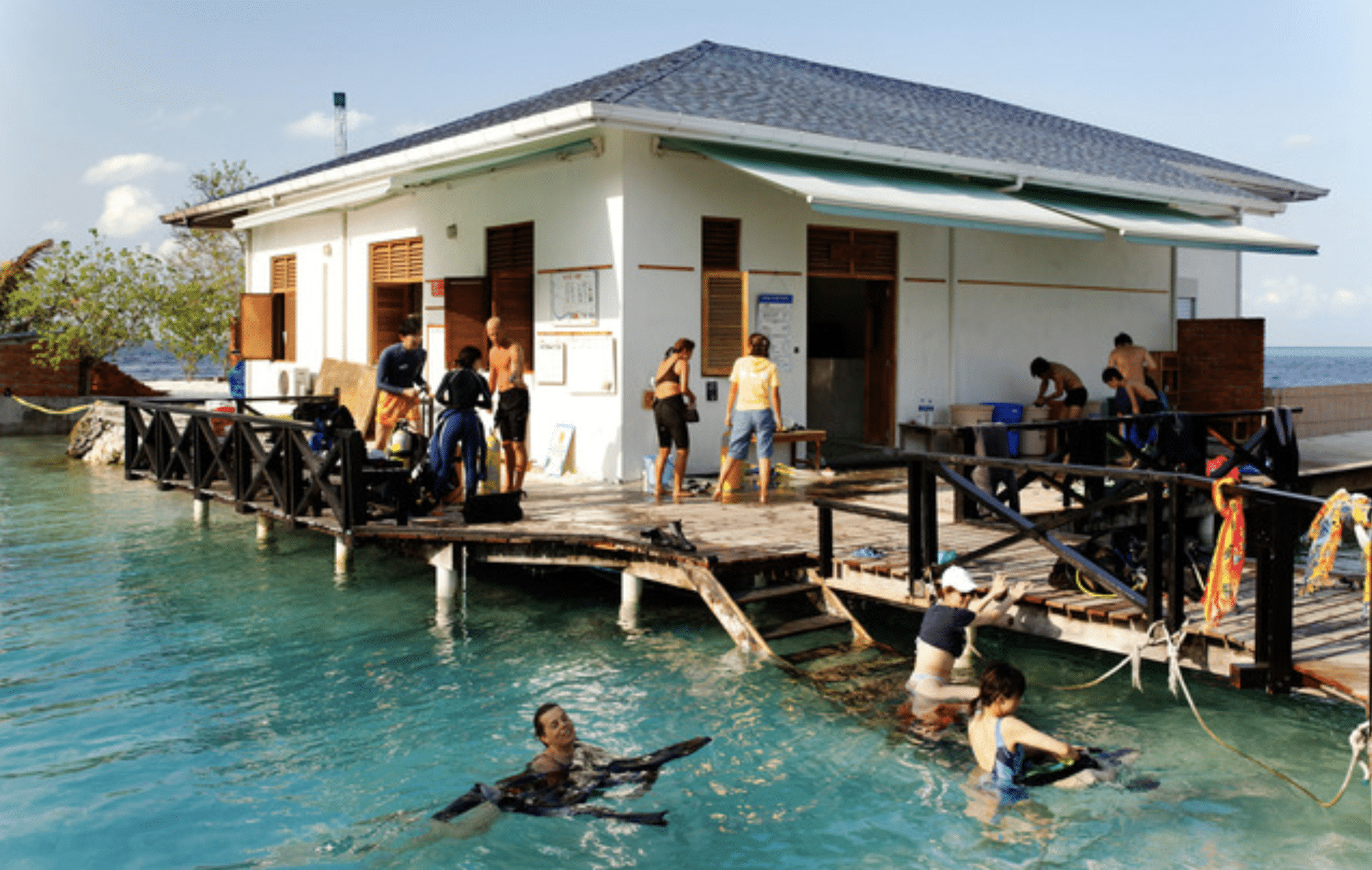 Screenshot 2026-04-07 at 002727 - N9BO℠ | Global Underwater Services Ltd People prepare for scuba diving at a waterside building with wooden walkways extending over clear blue water. Some are in wetsuits on the deck, while others are swimming or adjusting gear in the water.