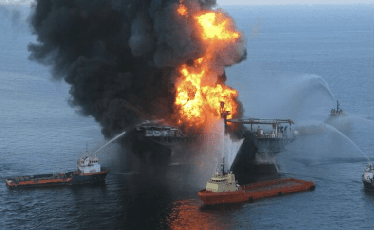 Several boats spray water to extinguish a large fire on an offshore oil platform, with thick black smoke and bright flames billowing into the sky over the sea.