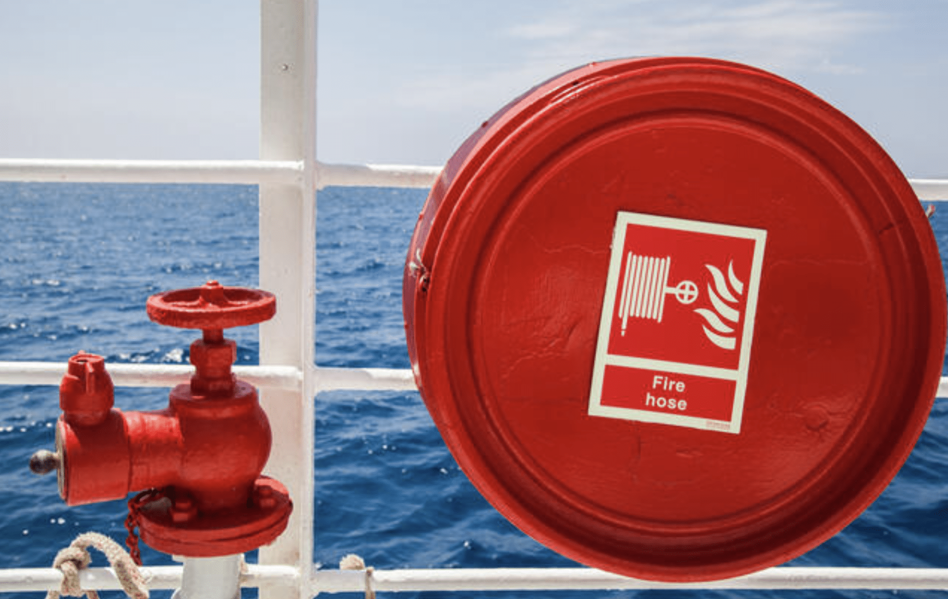 A red fire hose reel and valve mounted on a white metal rail of a ship, with the sea and blue sky visible in the background.