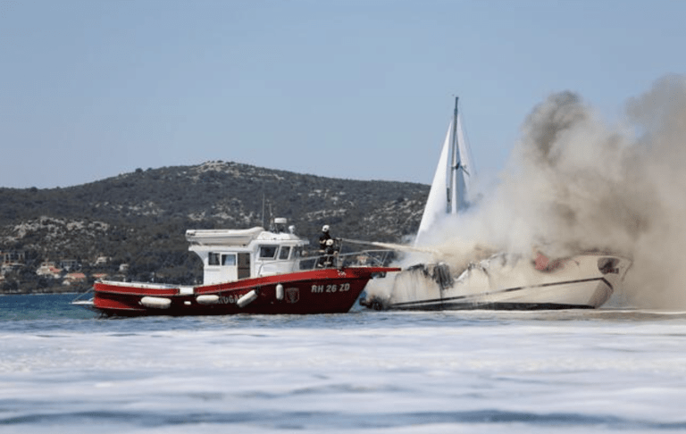 A red fire rescue boat sprays water on a burning white yacht emitting thick smoke, with green hills and buildings visible in the background.
