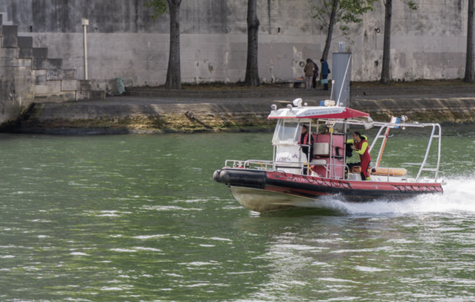 A small red and white motorboat with two people on board speeds along a green river, with a concrete embankment and a few trees visible in the background.