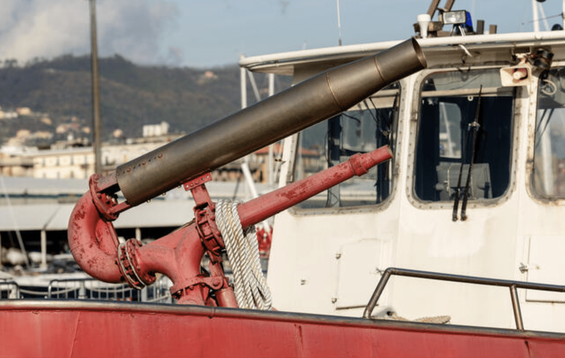 A close-up view of a large red water cannon mounted on the deck of a boat, with a white cabin and windows in the background and a hilly shoreline visible in the distance.
