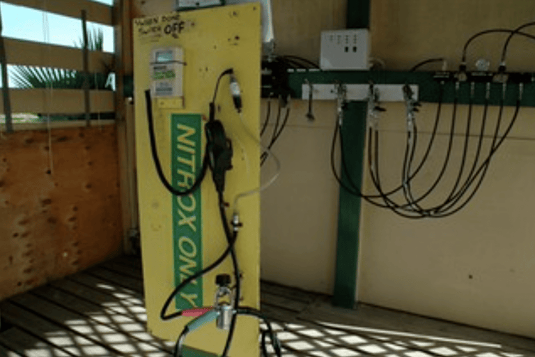 A yellow panel labelled NITROX ONLY is mounted vertically, featuring gauges, valves, and hoses, inside a shaded wooden structure with various tubes and a power switch visible on the wall.