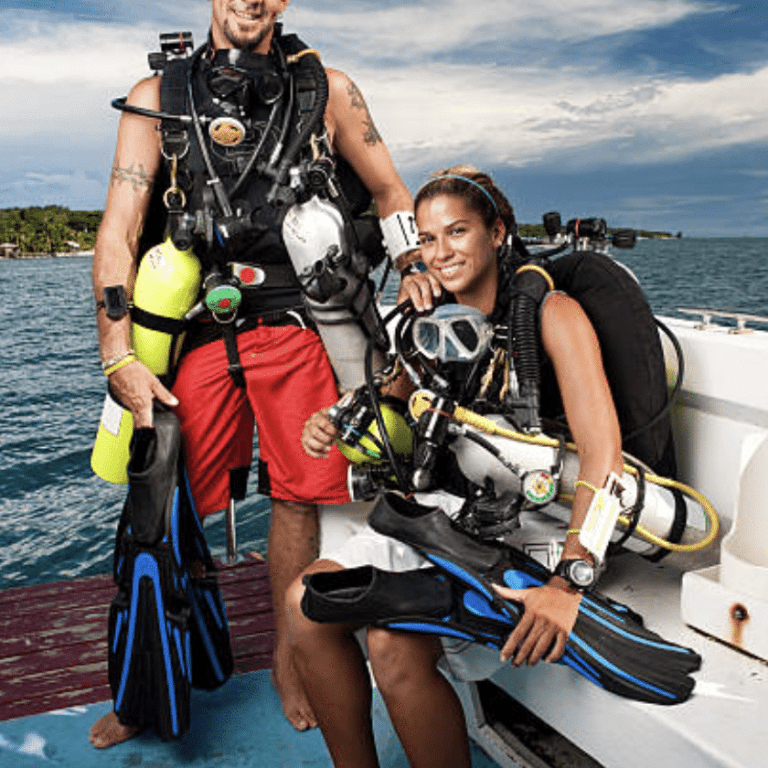 Two scuba divers in kit smile on a jetty next to a boat. One stands holding fins and cylinders, while the other sits with fins in hand. The sea and a partly cloudy sky are visible in the background.