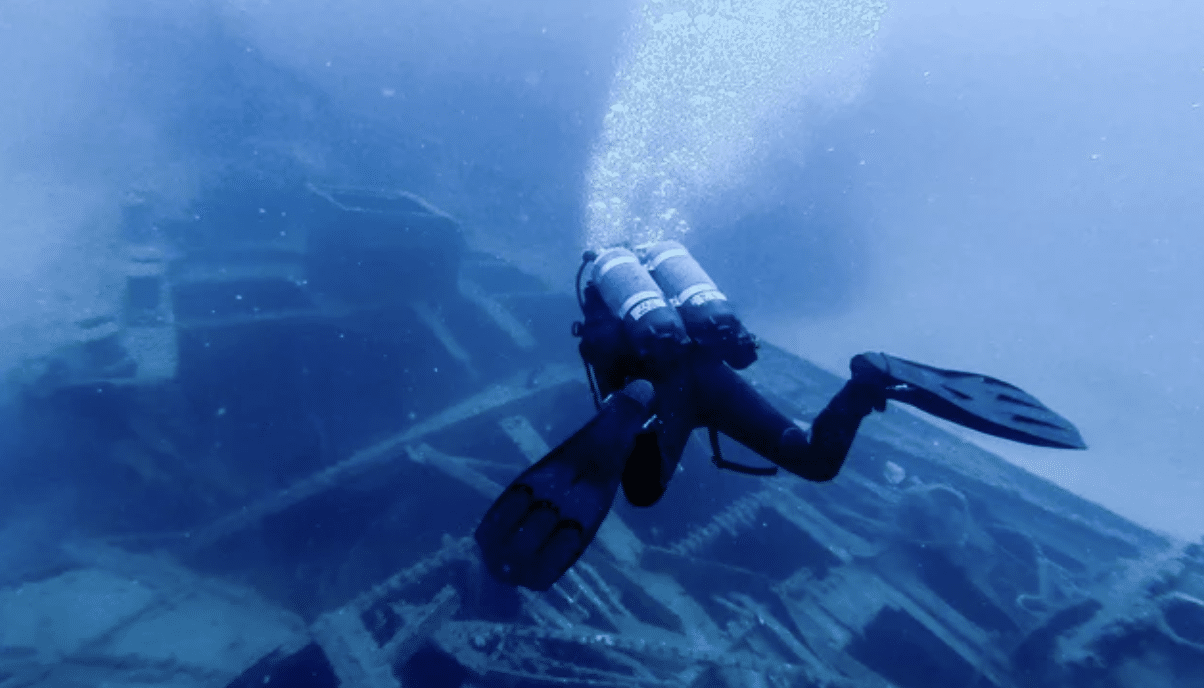 A scuba diver with twin cylinders and fins swims underwater near a large, sunken shipwreck, with bubbles rising to the water’s surface.