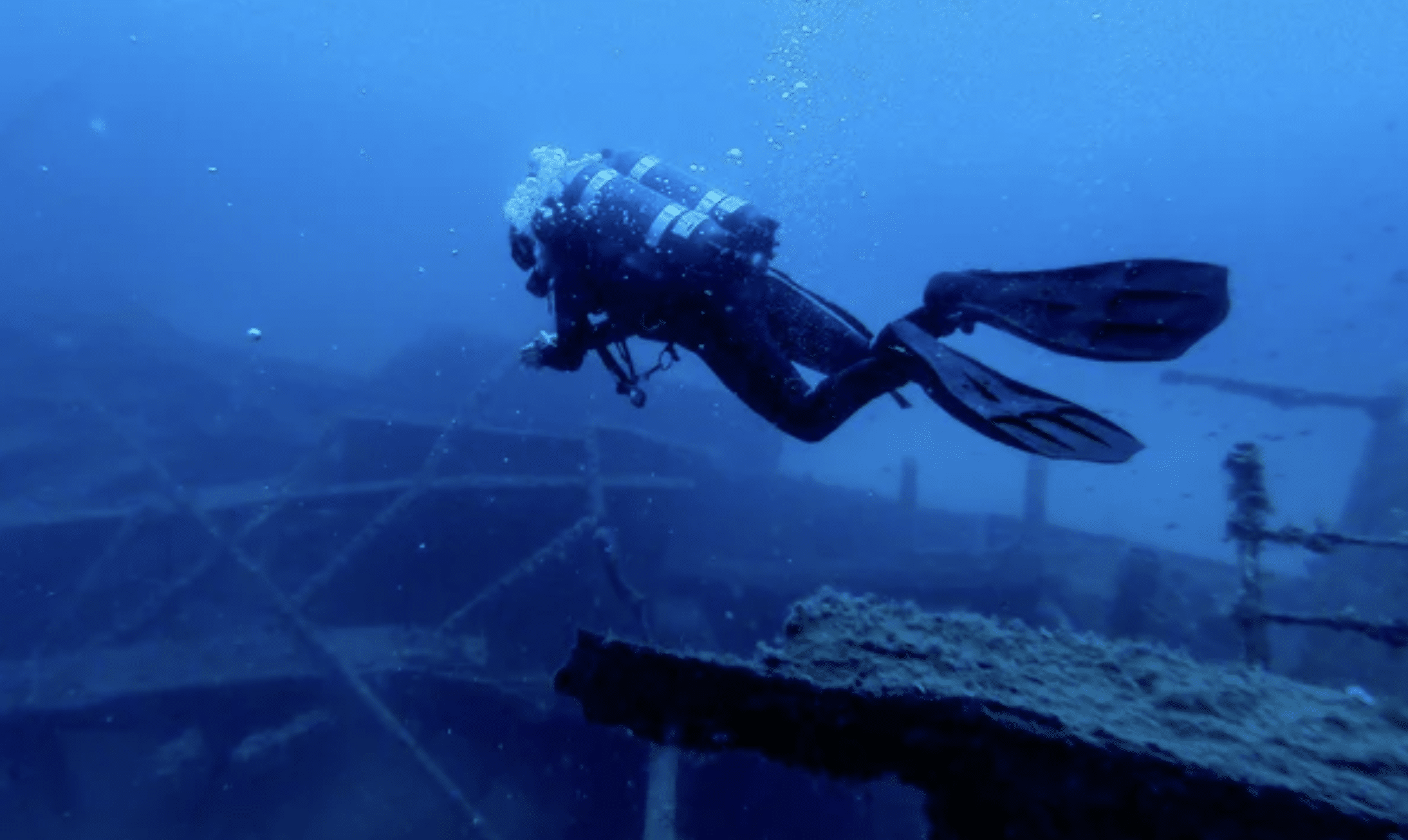 A scuba diver swims near the remains of a sunken shipwreck under deep blue water, surrounded by bubbles and scattered metal debris.