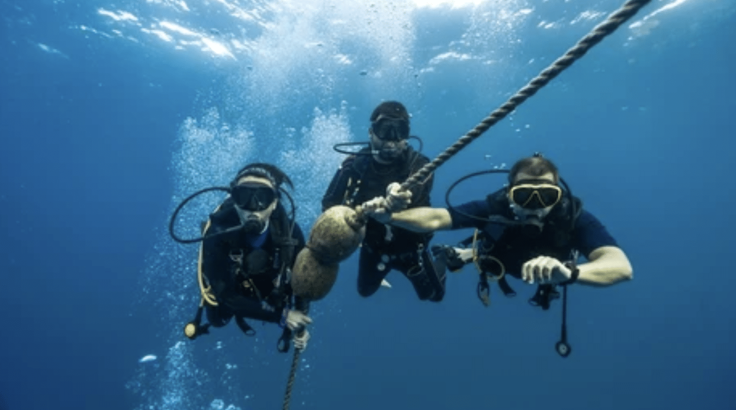 Screenshot 2026-04-15 at 221412 - N9BO℠ | Global Underwater Services Ltd Three scuba divers underwater hold onto a rope, wearing wetsuits, masks, and oxygen cylinders, surrounded by blue water and bubbles rising towards the surface.