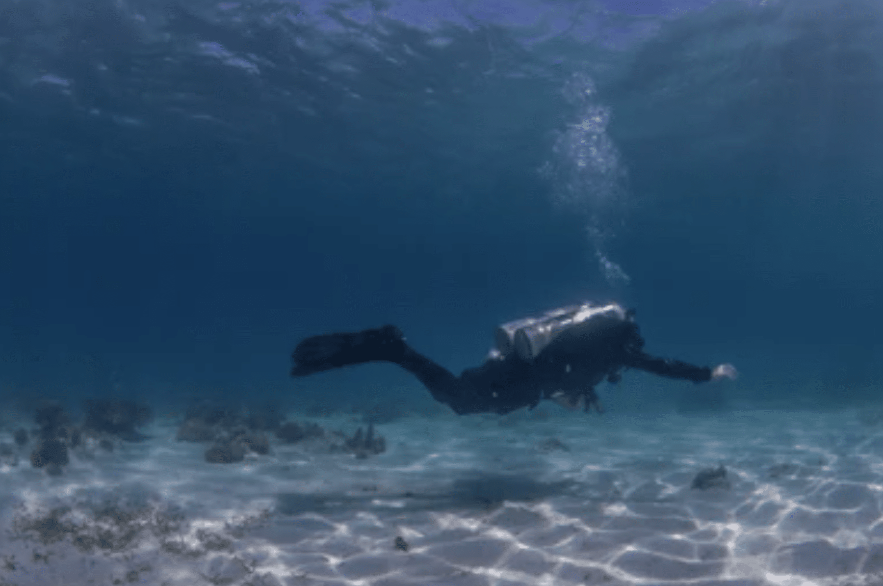 Screenshot 2026-04-15 at 221447 - N9BO℠ | Global Underwater Services Ltd A scuba diver swims underwater above a sandy seabed with patches of seaweed, surrounded by clear blue water and sunlight streaming from the surface.