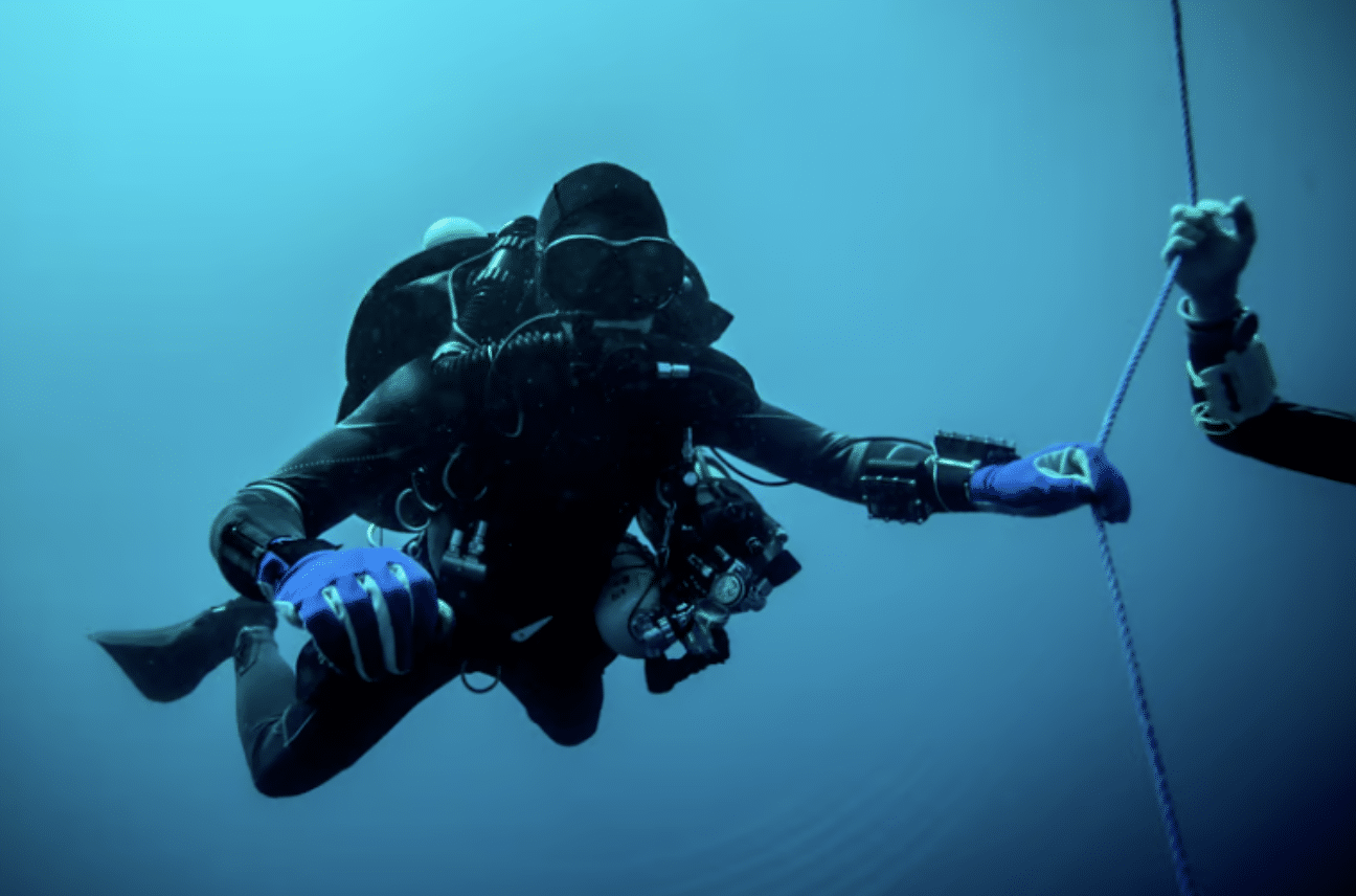 Screenshot 2026-04-15 at 233136 - N9BO℠ | Global Underwater Services Ltd A scuba diver in full kit, including gloves and mask, swims underwater while reaching towards a rope held by another diver, with a blue background indicating deep water.