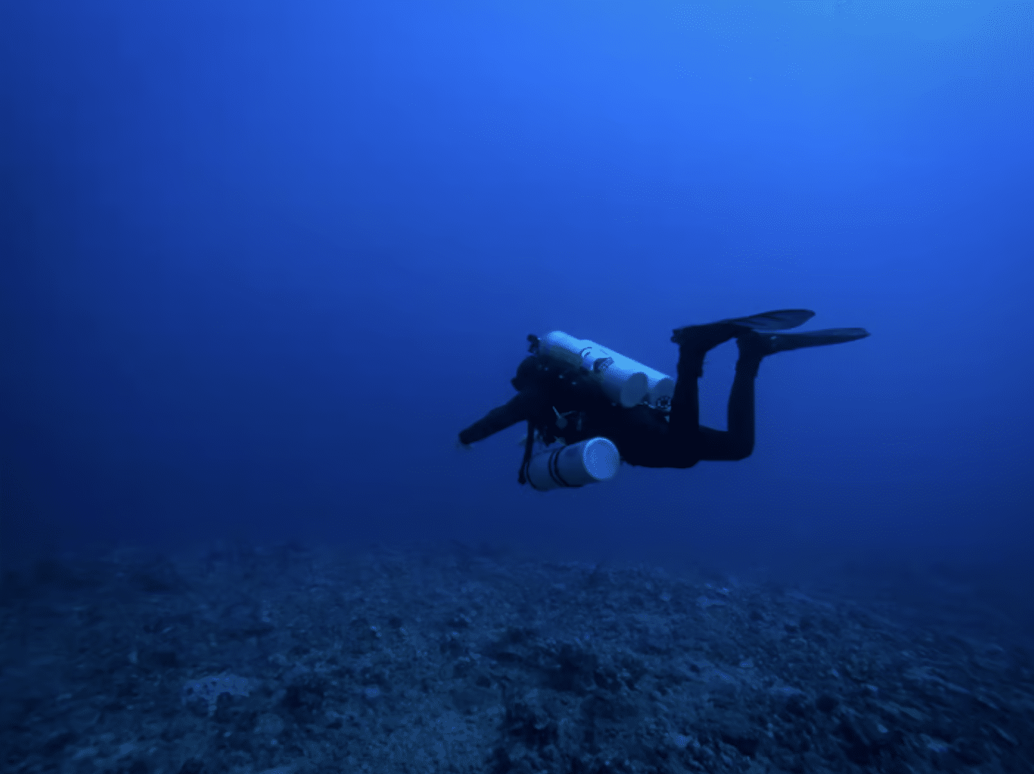 Screenshot 2026-04-15 at 233231 - N9BO℠ | Global Underwater Services Ltd A scuba diver with multiple cylinders swims near the sea floor in deep blue water, surrounded by an open, empty underwater environment.