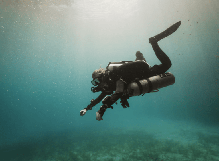A scuba diver in full kit floats underwater in clear blue-green water, surrounded by bubbles, with sunlight streaming from above. The diver appears to be exploring or examining the sea bed.