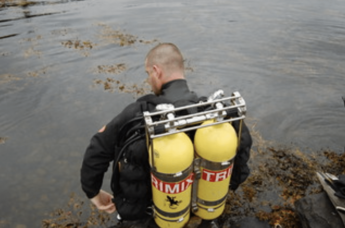 A scuba diver wearing a black wetsuit and carrying two large yellow oxygen cylinders on their back is entering the water, surrounded by seaweed and shallow rocks.