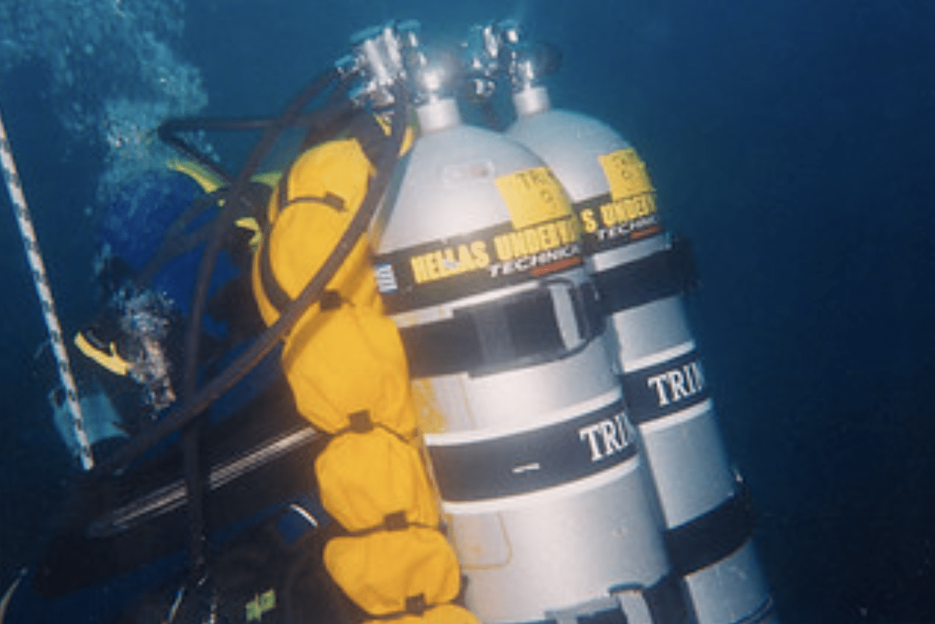 Screenshot 2026-04-16 at 011837 - N9BO℠ | Global Underwater Services Ltd A scuba diver underwater is carrying large oxygen cylinders labelled “TRIMIX” and “HELLAS UNDERWATER TECHNICAL,” with yellow equipment attached to the cylinders by hoses.