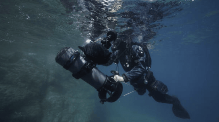 A scuba diver in full kit navigates underwater, holding onto an underwater scooter. The water is clear, and the diver appears focused as they move near submerged rocks.
