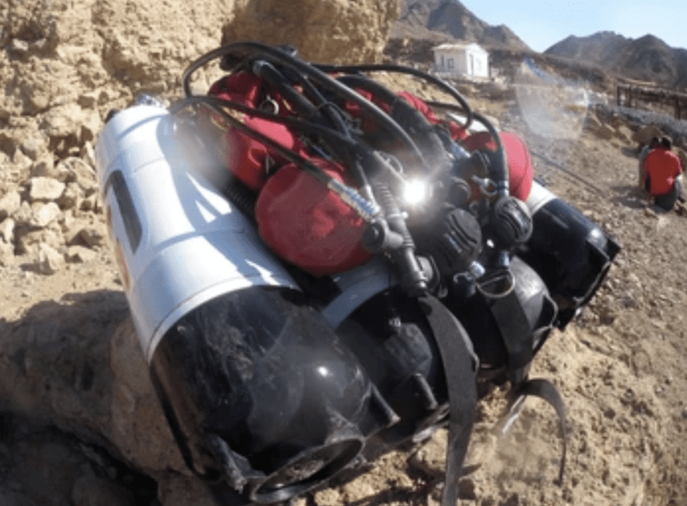 A set of scuba diving tanks and equipment, including multiple cylinders and hoses, rests on rocky terrain with mountains and a building visible in the background.
