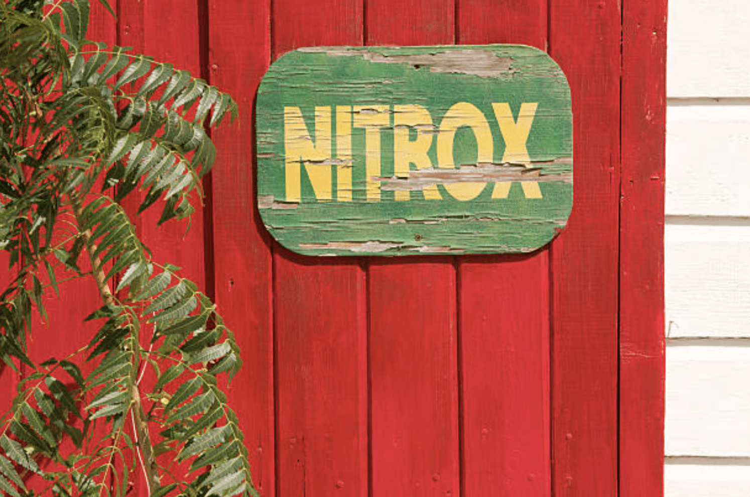 A weathered green sign with yellow NITROX text hangs on a red wooden wall, with green leafy branches partially visible on the left side.