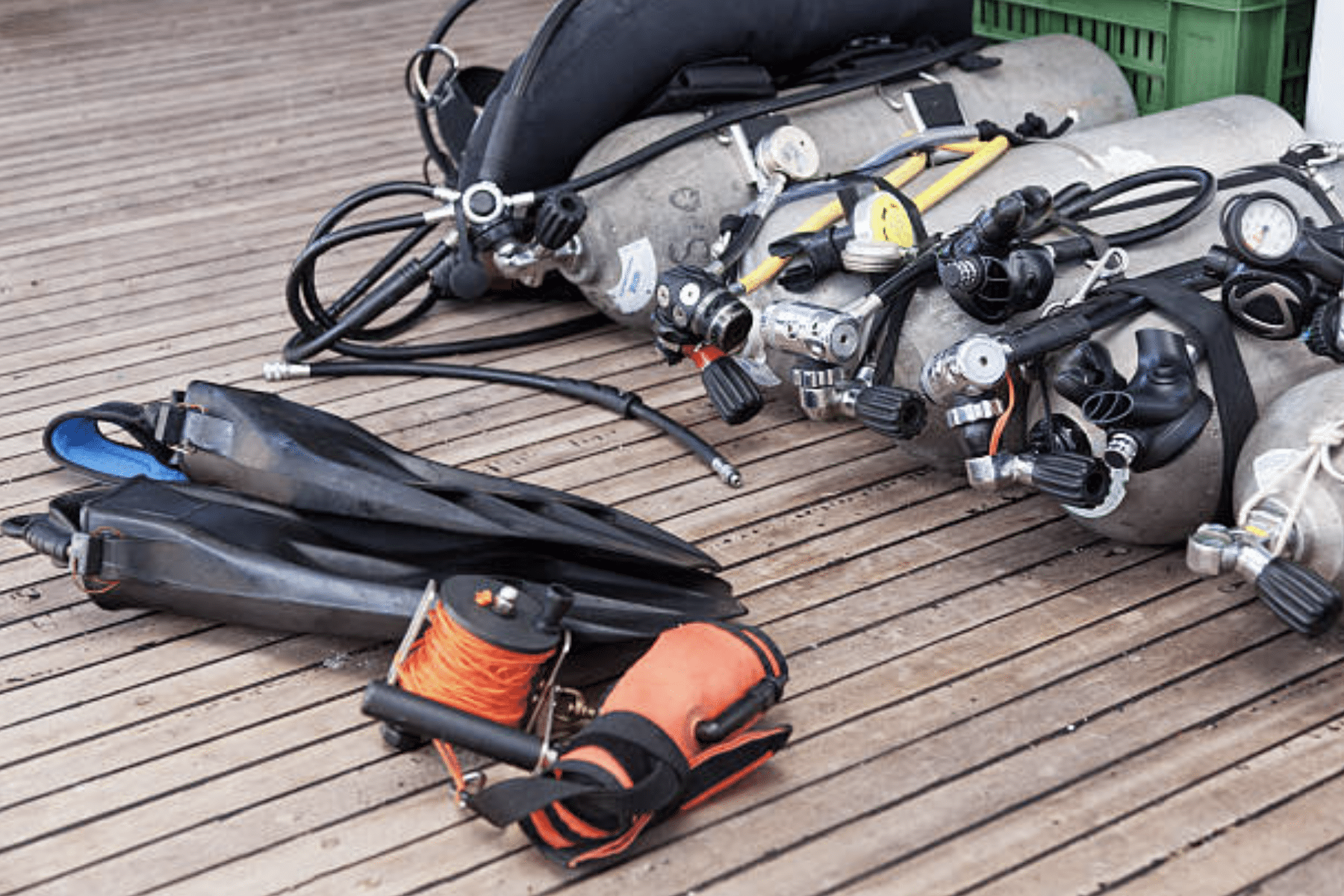 Several scuba diving cylinders with attached regulators lie on a wooden deck beside pairs of fins and an orange dive reel. The equipment appears organised and ready for use.