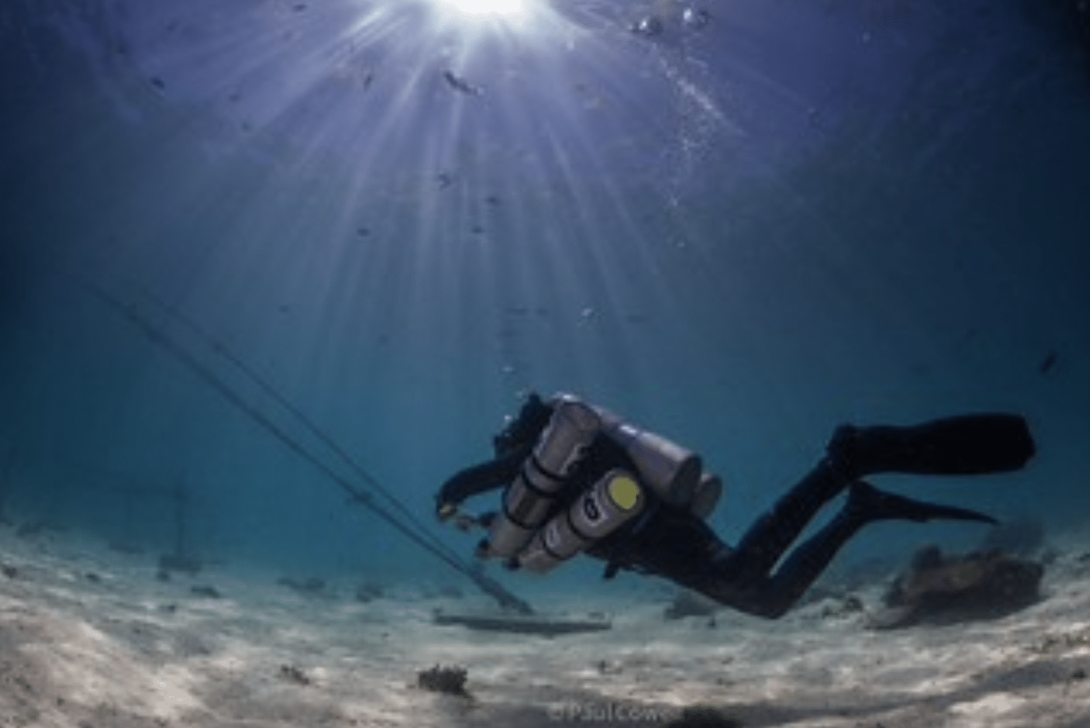 A scuba diver swims near the sandy sea floor under rays of sunlight, surrounded by clear blue water and some scattered aquatic plants and equipment.
