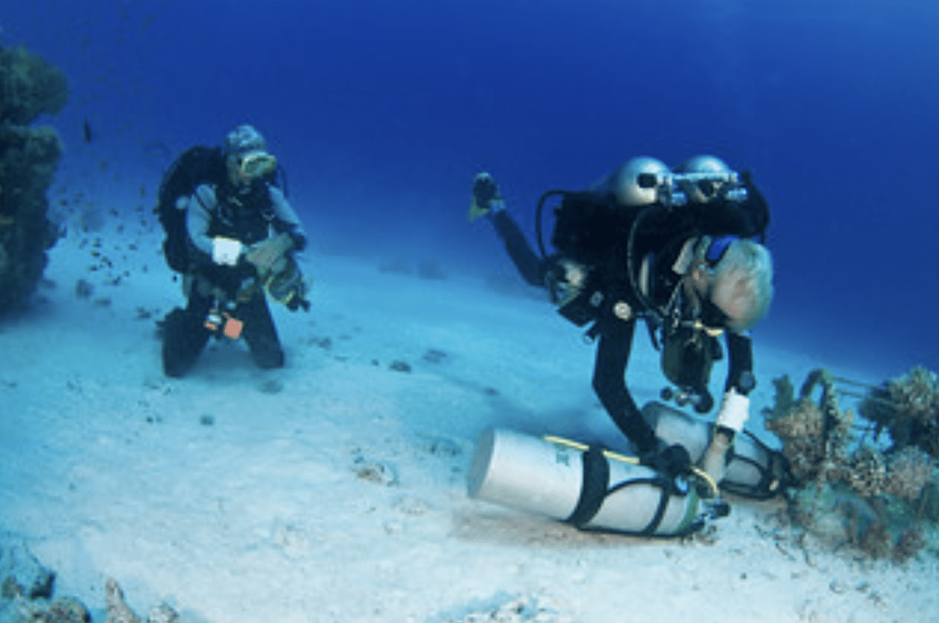 Screenshot 2026-04-19 at 181627 - N9BO℠ | Global Underwater Services Ltd Two scuba divers in full kit are underwater near the sea floor. One diver is placing or inspecting a cylinder on the sandy seabed, whilst the other observes nearby with coral and small fish in the background.
